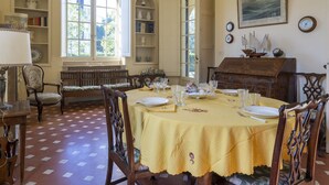 A dining room with a table set for four, surrounded by wooden chairs. There are bookshelves with decorative items and a piano in the background.