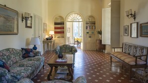 A spacious living room with a floral sofa, a wooden bench, and a coffee table. There are bookshelves and a dining area in the background.