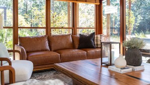 Un salon chaleureux avec un canapé en cuir marron, une table basse en bois et une vue sur une forêt, visible à travers de grandes fenêtres.