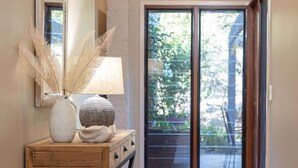 Un couloir avec une console en bois, une lampe, des objets décoratifs, un panier en osier et, à travers une porte vitrée, une vue sur un espace extérieur.