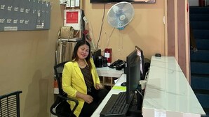 Una mujer sentada en un escritorio con una computadora, un televisor montado en la pared y un ventilador.