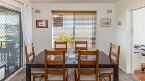Un coin repas avec une table et des chaises en bois, une fenêtre avec des stores et une porte dotée d’un portail en métal.