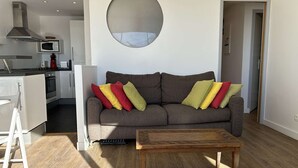 A modern living room with a grey sofa, colorful cushions, a wooden coffee table, and a kitchen area with a microwave and stove. - Air-conditioned Apartment 6 pax with Terrace and Pool in Port Camargue (Port Camargue)