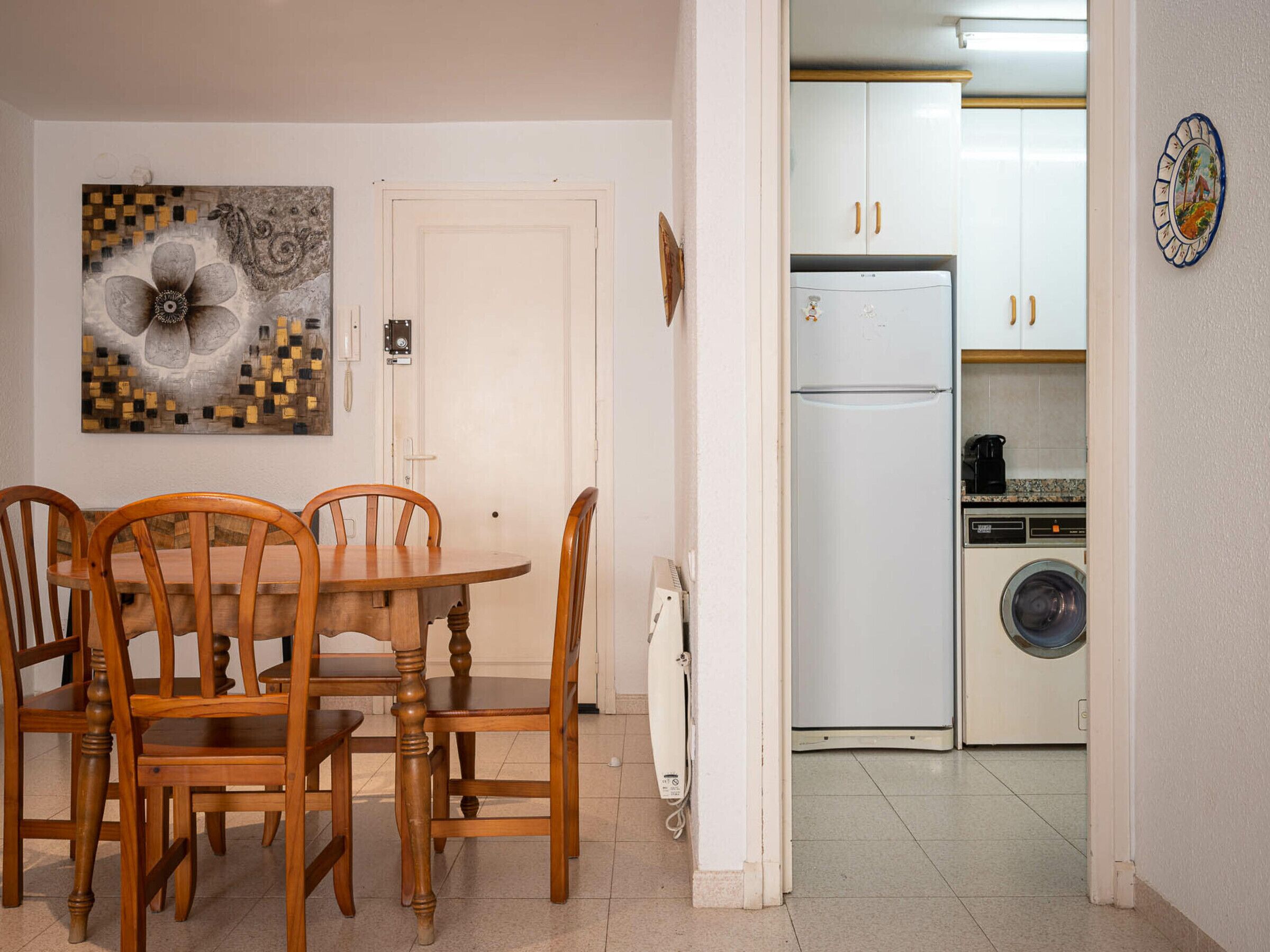 A dining area with a wooden table and chairs, a white door, and a kitchen with white cabinets and appliances.