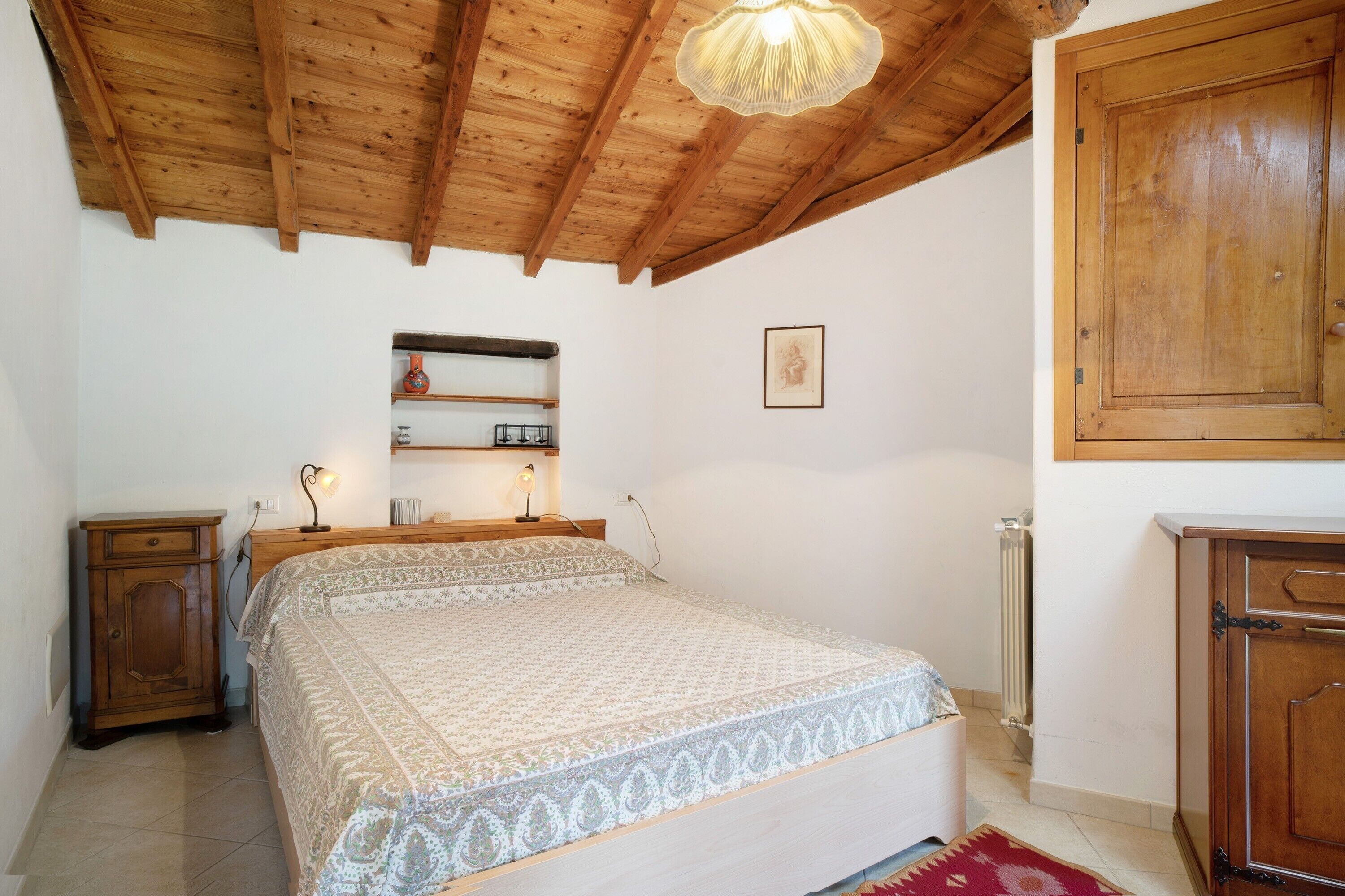 Un dormitorio con techo de madera, una cama con ropa de cama estampada, una mesita de noche, un aparador y un radiador.