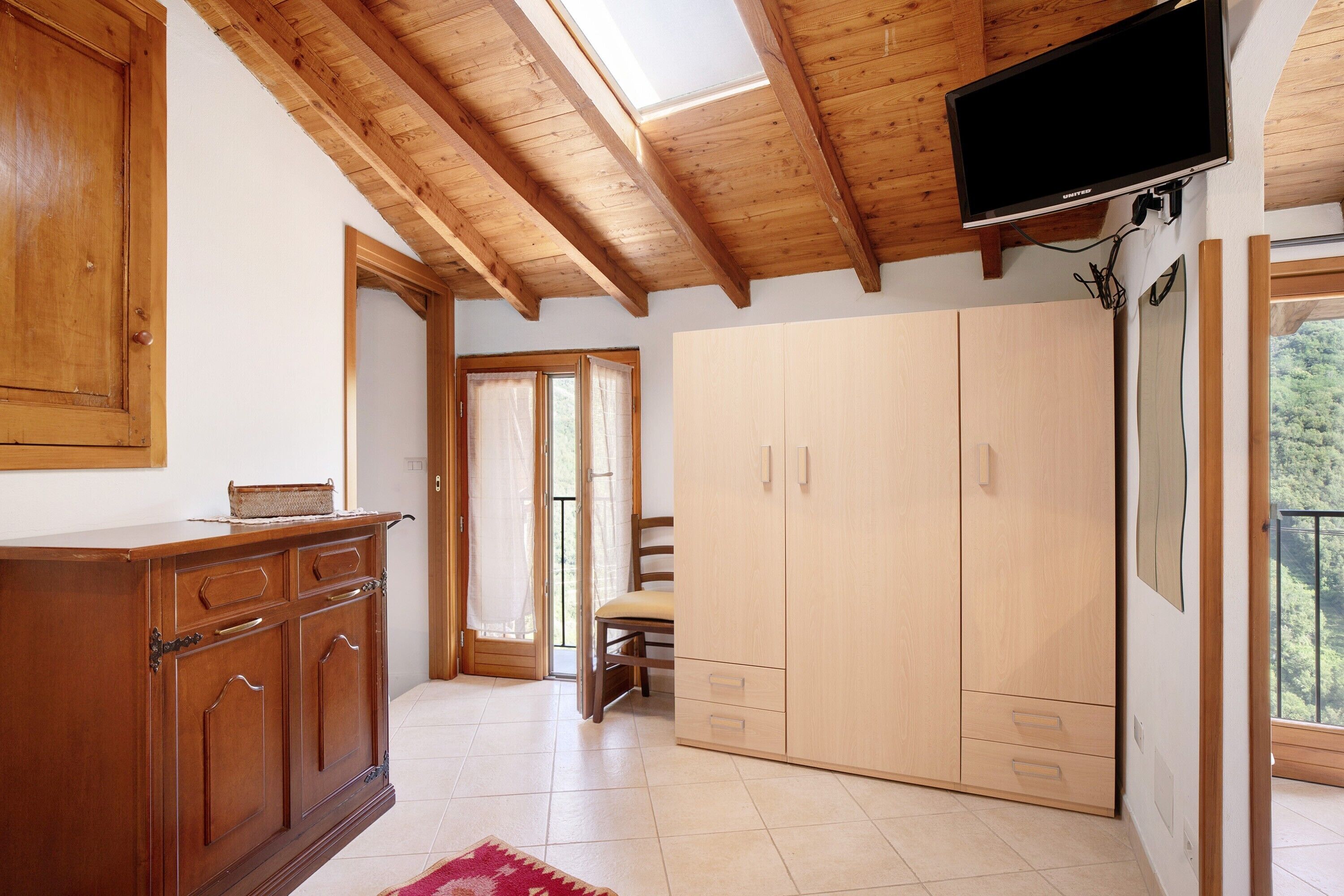 A room with wooden ceiling beams, a TV mounted on the wall, a wooden wardrobe, and a chair near a door.