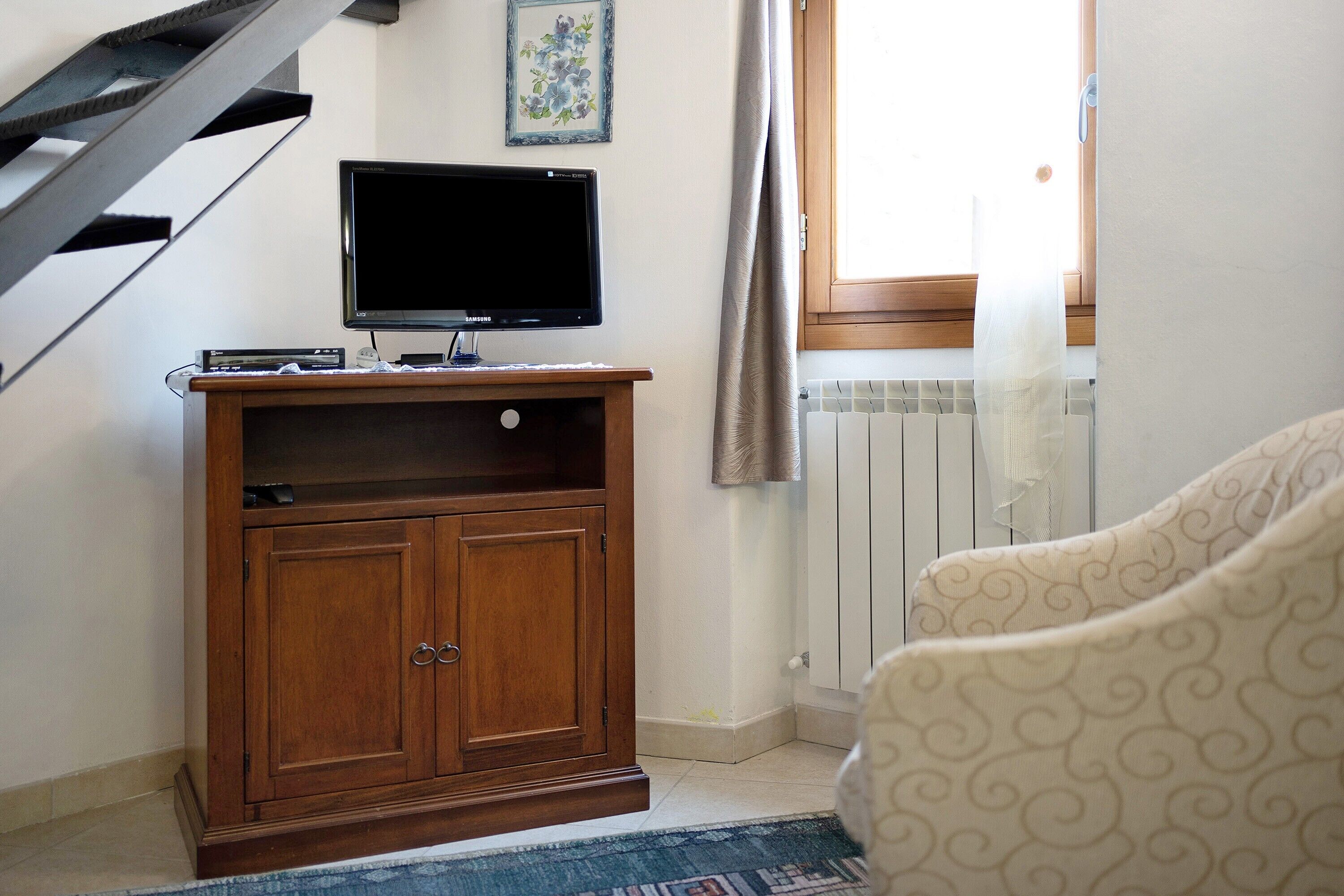 Una habitación con un mueble de entretenimiento de madera, un televisor, un sillón estampado, una escalera y una ventana con cortinas.