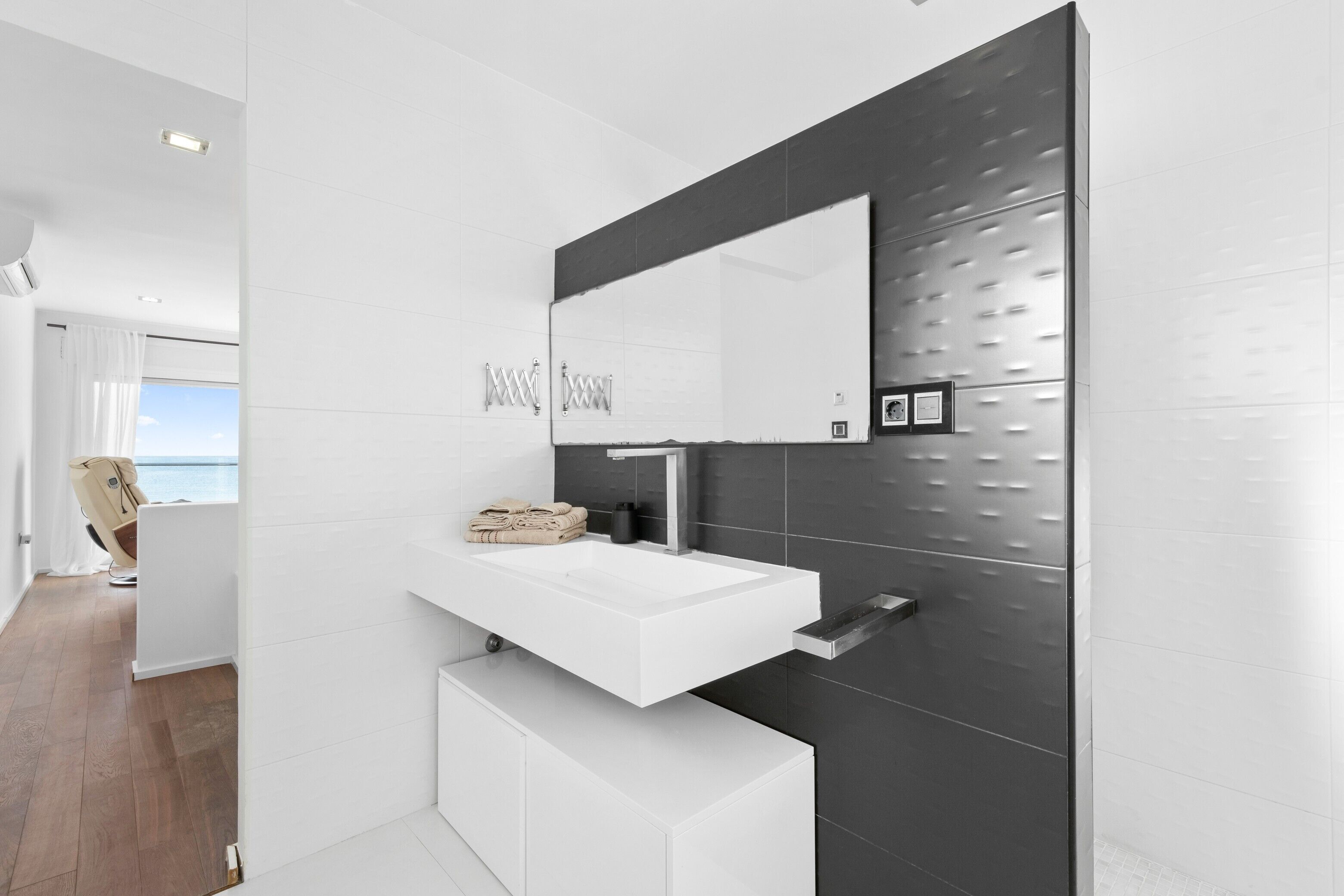 A modern bathroom with a white vanity, a large mirror, and a view of the sea.