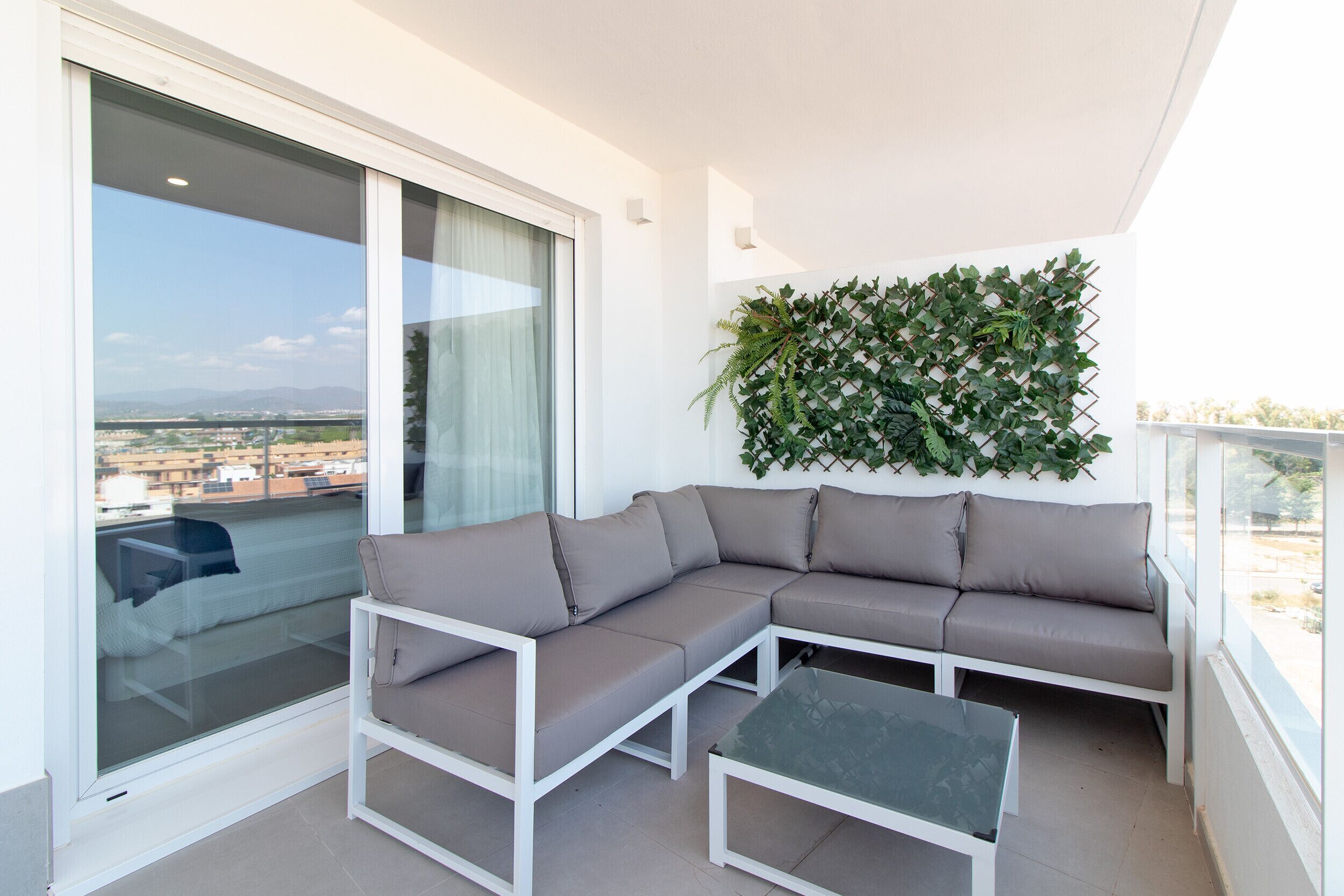 Ein Balkon mit einer Eckcouch und einem Glastisch, ein großes Fenster mit Blick auf Gebäude und eine grüne Pflanzenwand.