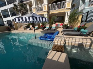 Un área de piscina con sillas de descanso, una sombrilla a rayas y plantas en macetas.