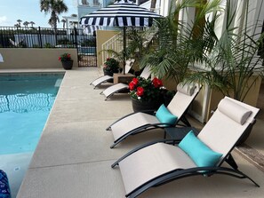 Uma área à beira da piscina com espreguiçadeiras, um guarda-sol listrado e plantas em vasos.