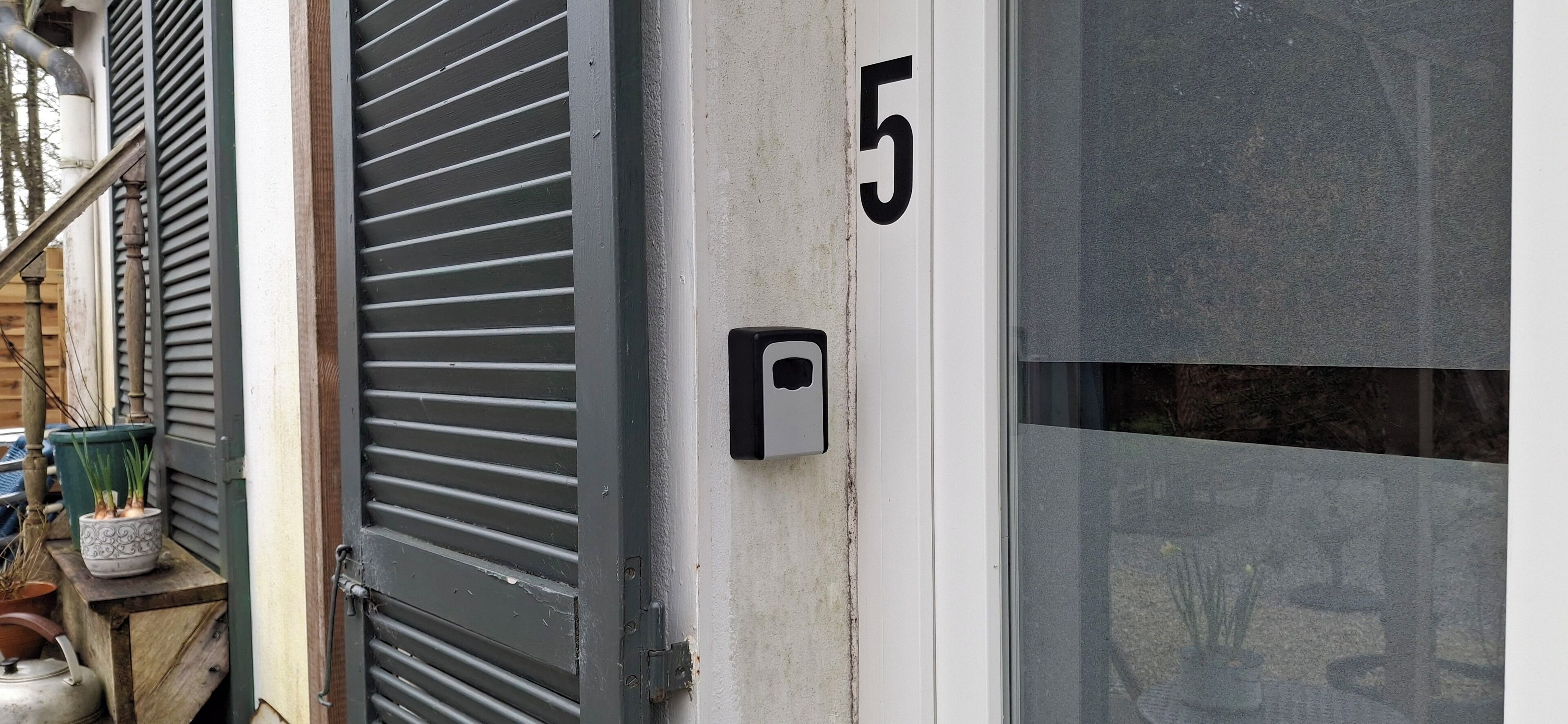 Une porte avec une plaque portant le numéro « 5 », une boîte aux lettres et un pot de fleurs posé sur une étagère en bois.