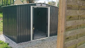 Un abri de jardin en métal avec une porte, une base en gravier et une clôture en bois en arrière-plan.