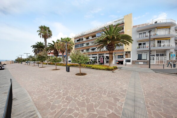 A paved walkway with palm trees, a multi-story building with balconies, and a clear sky.