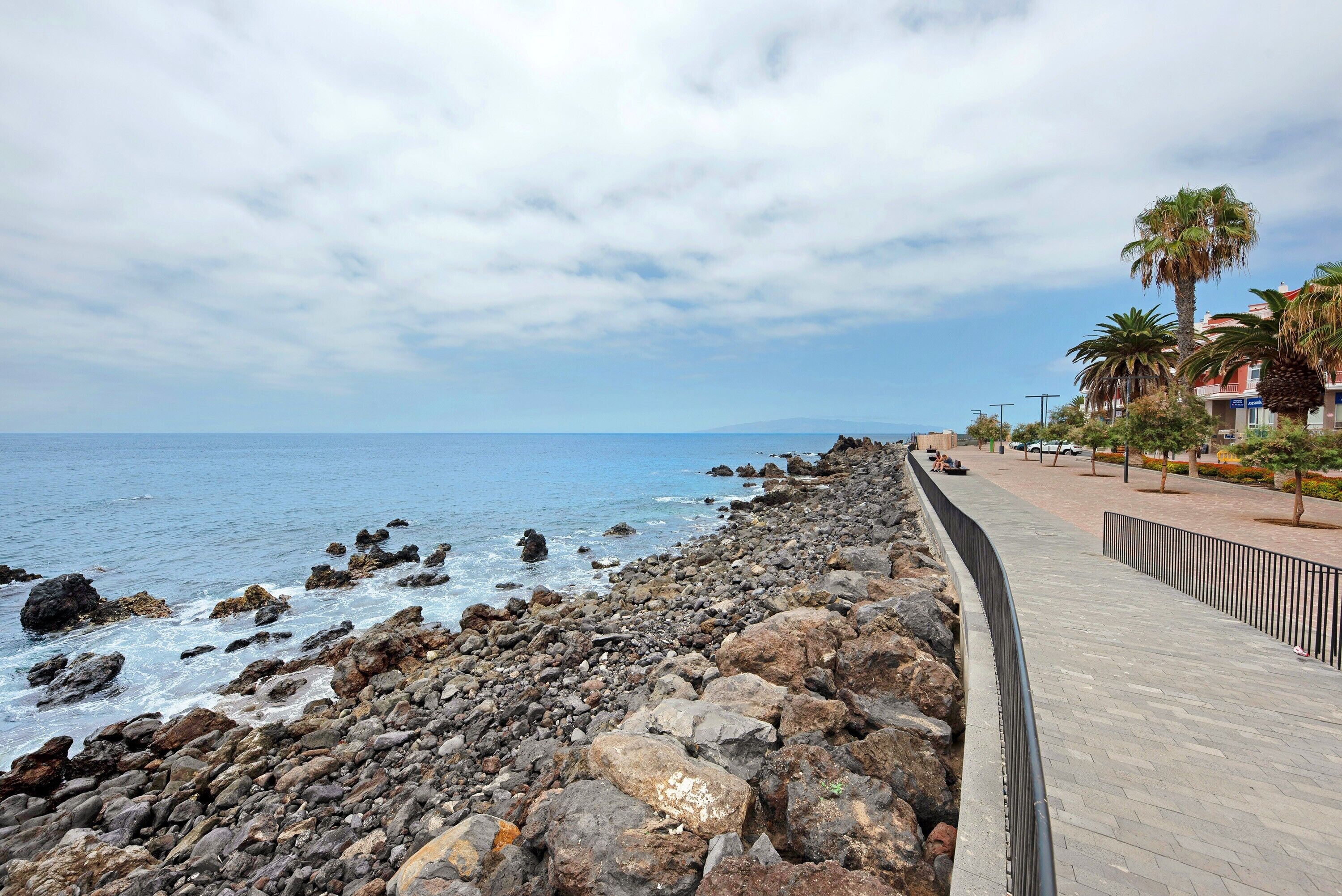 Un paseo marítimo costero con una línea de costa rocosa, un sendero pavimentado y palmeras.