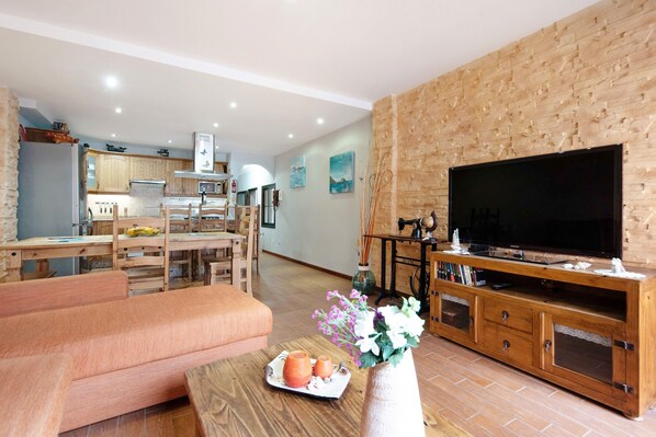 A modern living room with a sofa, a coffee table with decorative items, a wooden entertainment unit with a TV, and a kitchen in the background.