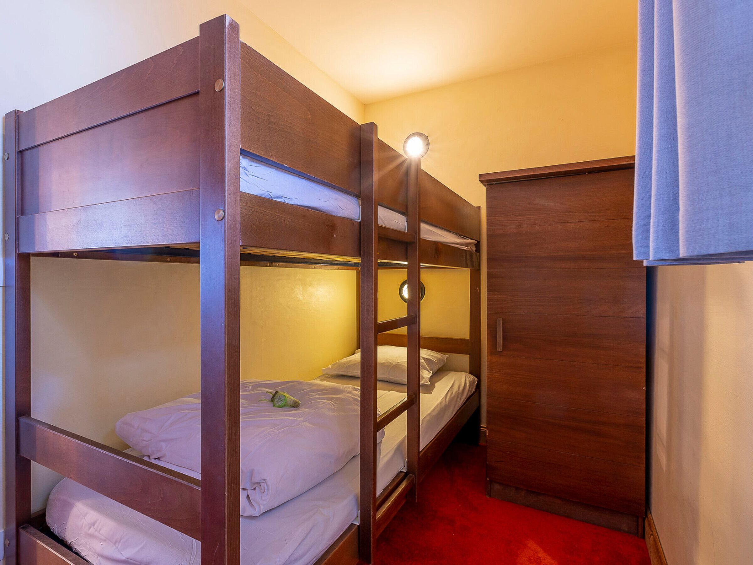 A bunk bed room with a wooden bunk bed, a small cabinet, and a red carpet.