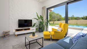 Una sala de estar moderna con un televisor de pantalla plana, un sofá, un sillón amarillo, una mesa de centro y un ventanal con vistas a una piscina.