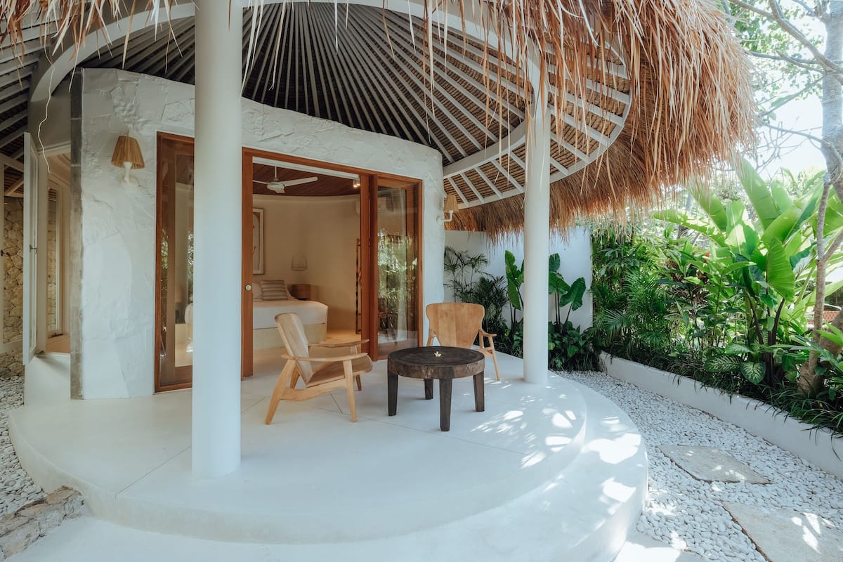 A covered outdoor seating area with wooden chairs and a table, surrounded by white walls and a thatched roof.