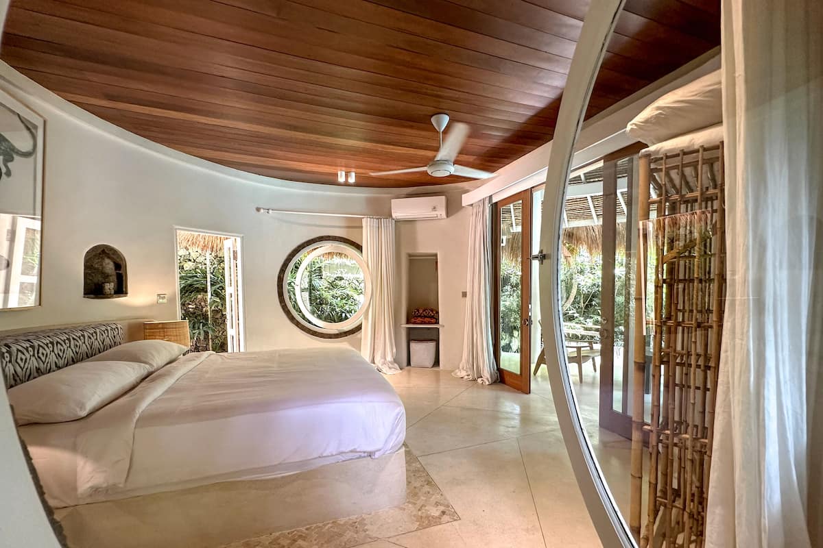 A bedroom with a bed, a ceiling fan, a window with a view of greenery, and a sliding glass door leading to an outdoor area.