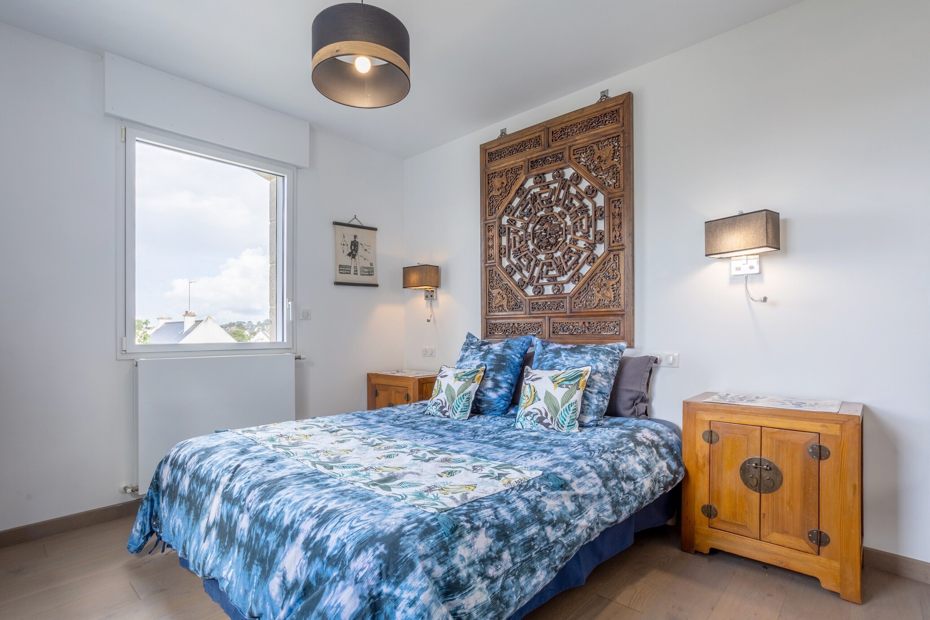 A bedroom with a large bed, a window, a wall tapestry, bedside lamps, and a wooden cabinet.
