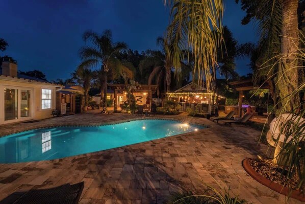 A swimming pool with a tiled deck, palm trees, and a gazebo with string lights.