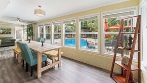 A sunlit room with a wooden dining table, chairs, and a view of a swimming pool.