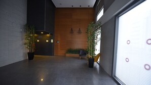 A modern lobby with a dark floor, wooden wall, and a large window with blinds. There are potted plants and a bench. - The apartment in the hitech park Haifa (Haifa)