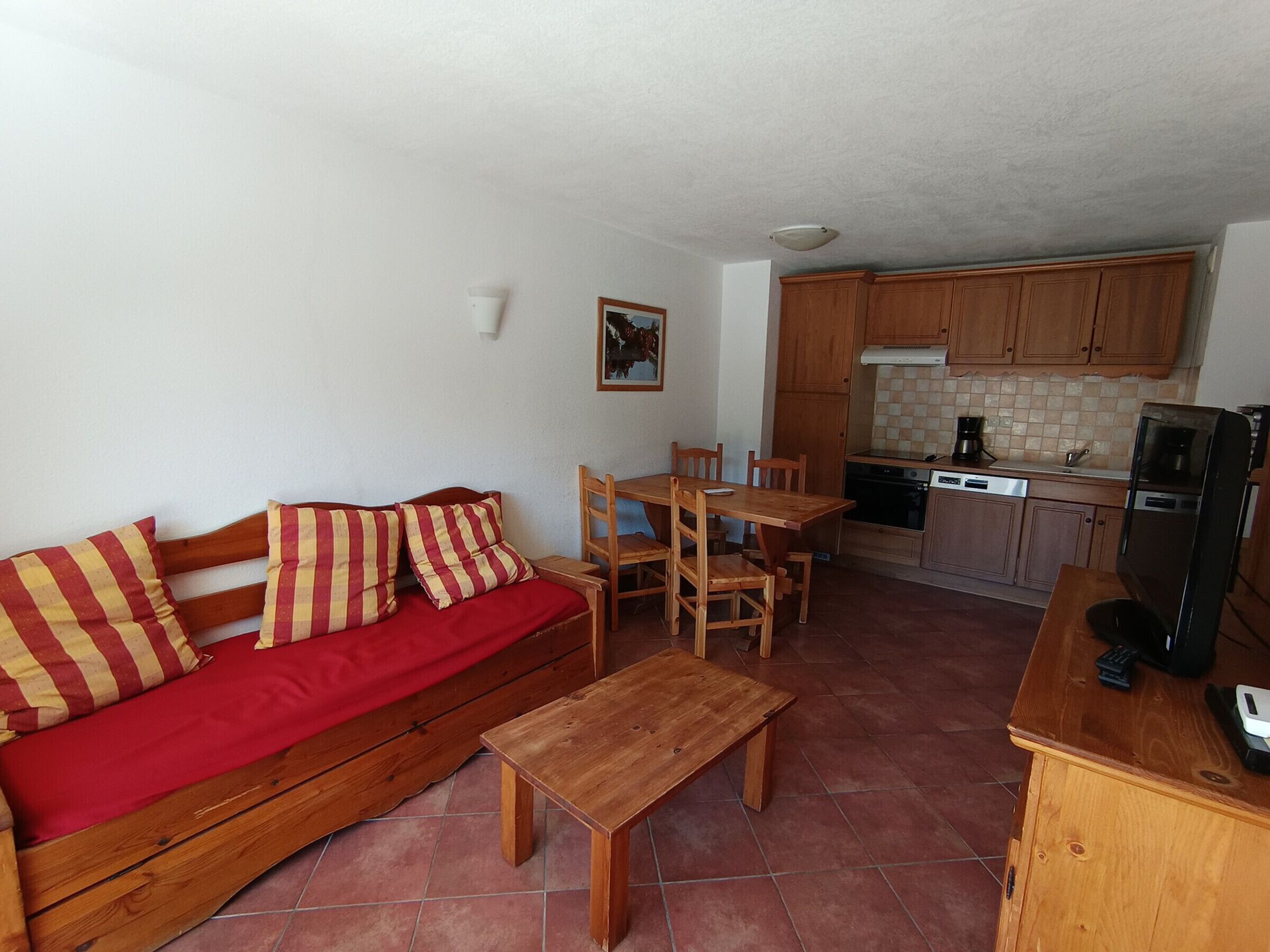 A compact living space with a wooden sofa, a small wooden table, a dining table with chairs, a kitchen area with wooden cabinets, and a tiled floor.