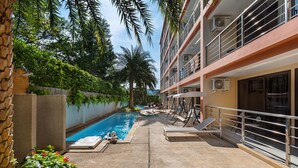 A pool area with lounge chairs and umbrellas, surrounded by greenery and a building with balconies. - Rawai Beach Condo Room 407 (Rawai)
