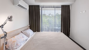 A hotel room with a bed, a desk lamp, an air conditioner, and a view of the outdoors through large windows. - Rawai Beach Condo Room 407 (Rawai)
