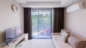 A modern living room with a flat-screen TV on a wooden stand, a sofa with cushions, a large window with curtains, and an air conditioning unit on the wall. - Rawai Beach Condo Room 407 (Rawai)