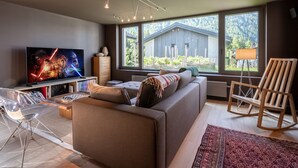 A modern living room with a large TV, a sofa, a wooden chair, and a view of a house through large windows.