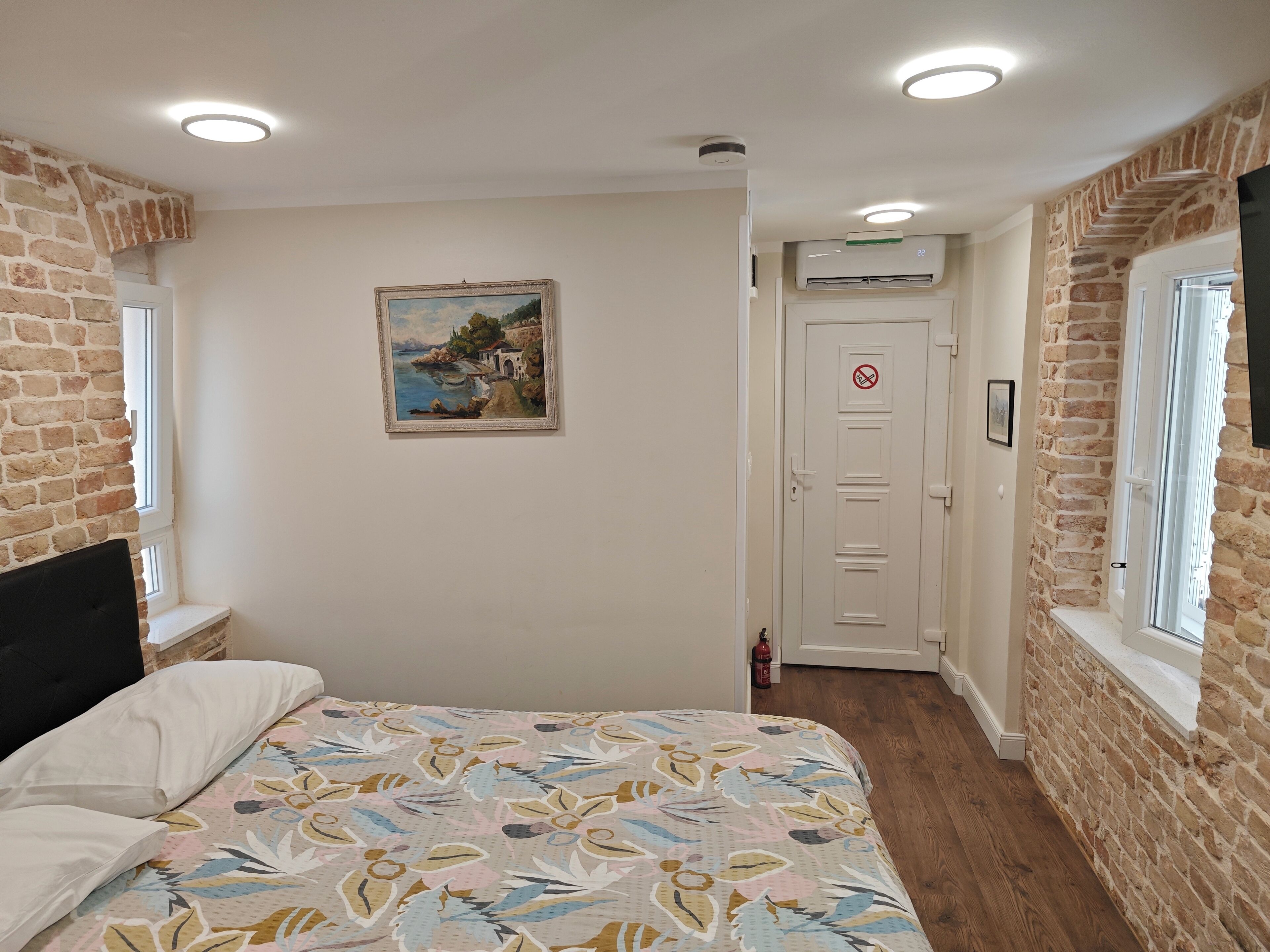 Quarto com uma cama, um quadro, uma porta com placa de proibido fumar e paredes de tijolo.