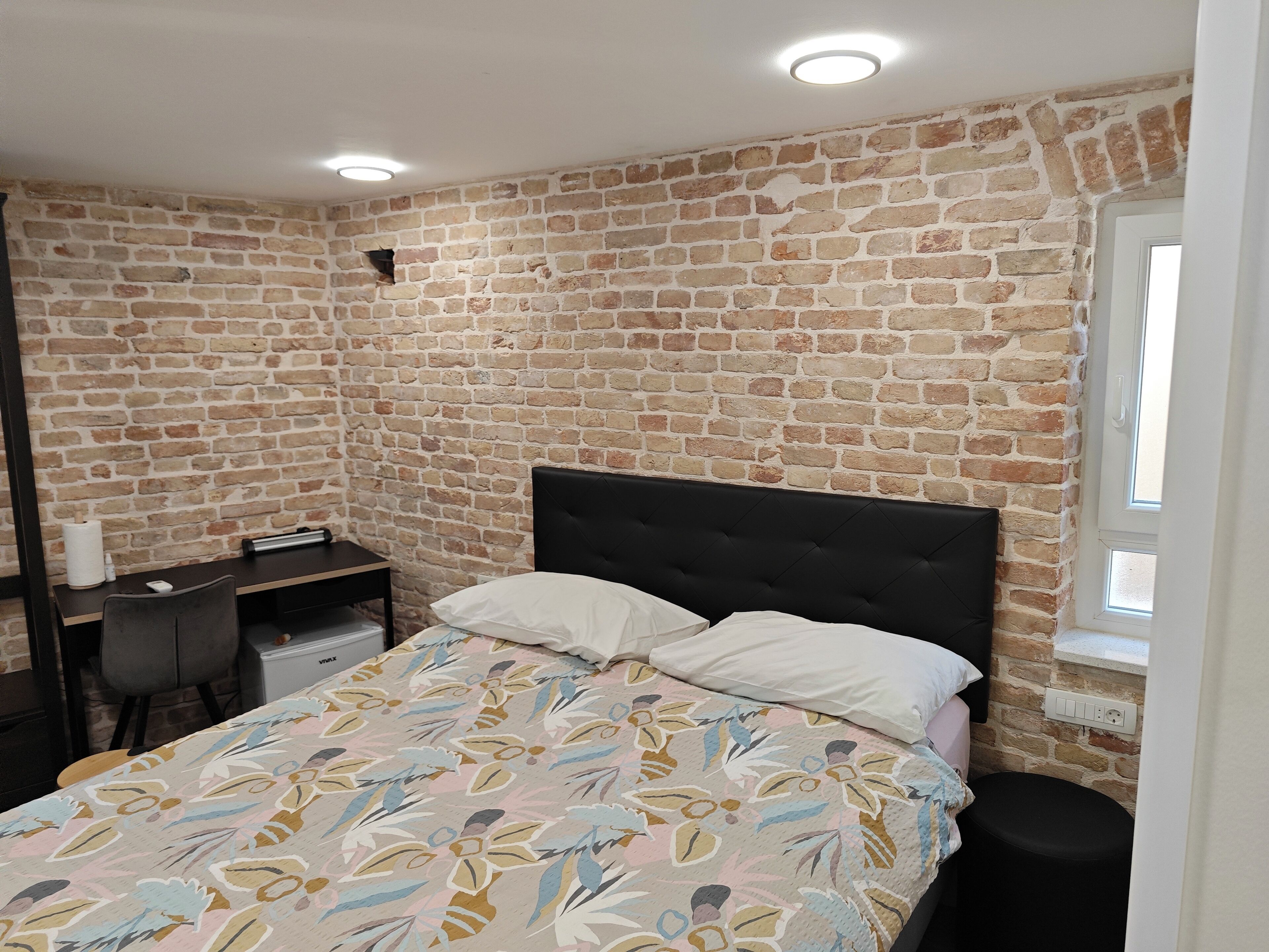 A bedroom with a brick wall, a bed with floral bedding, a desk, and a window.