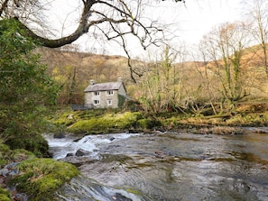 Une maison en pierre, avec une rivière devant, entourée d’arbres et de verdure.