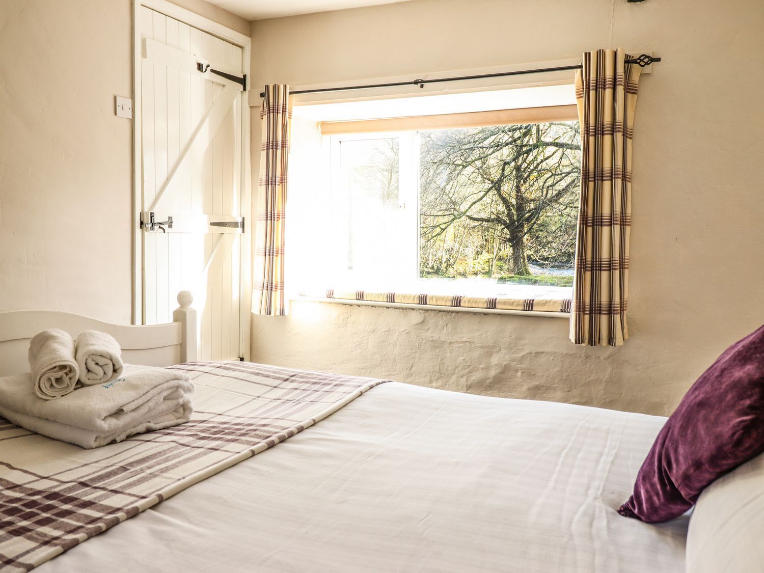 A bedroom with a bed, a window with curtains, a bedside table with towels, and a view of trees outside.