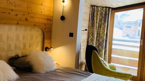 Une pièce chaleureuse avec une tête de lit en bois, un lit avec des oreillers, une table de chevet avec une lampe et une vue sur un bâtiment à travers une fenêtre ornée de rideaux à motifs.
