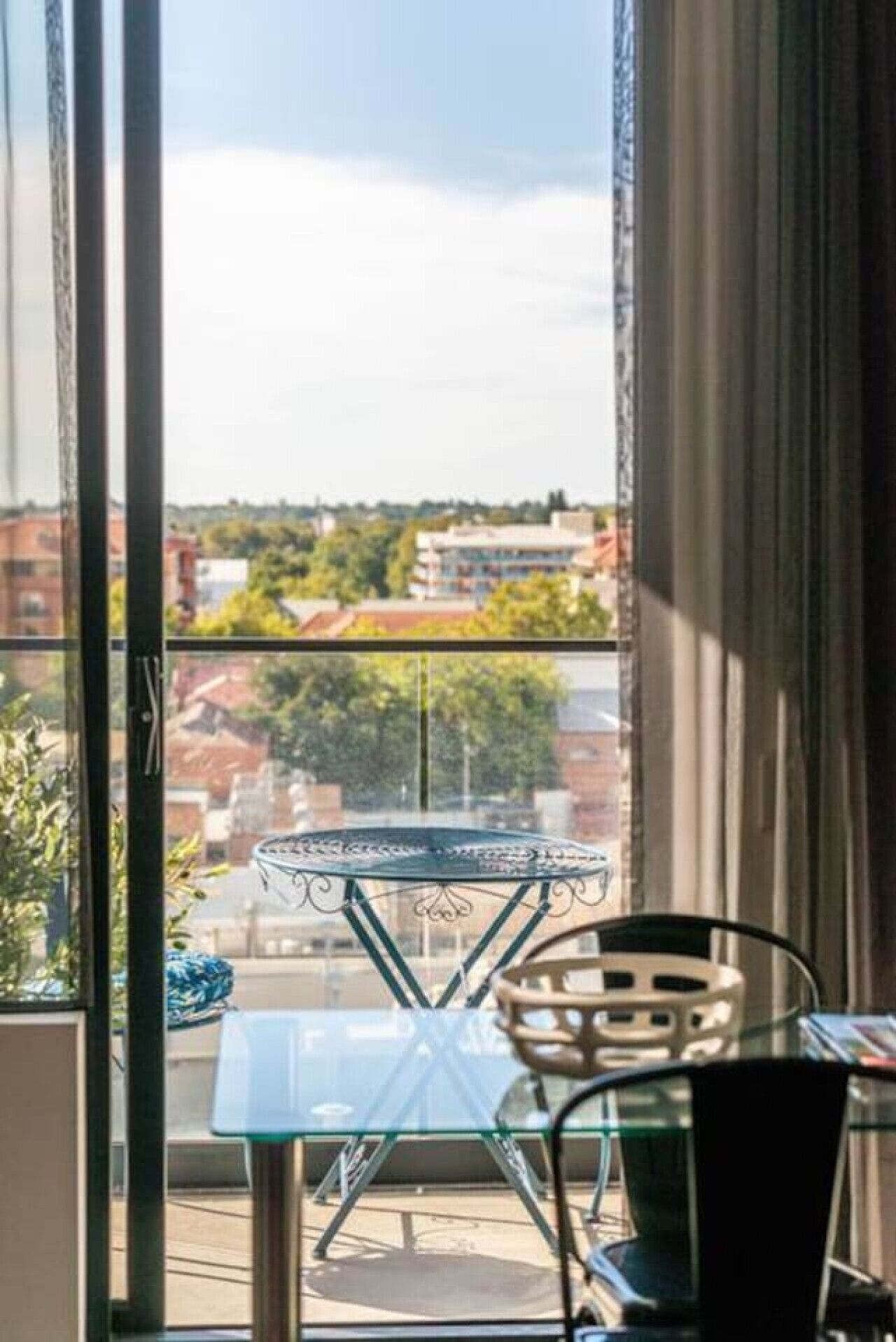 En balkong med ett glasbord och stolar, med utsikt över en stadsmiljö med byggnader och träd.