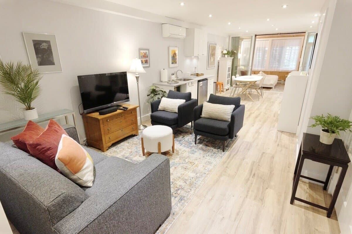 Een moderne woonkamer met een bank, fauteuils, een televisie, een bijzettafeltje en een keuken in de achtergrond.