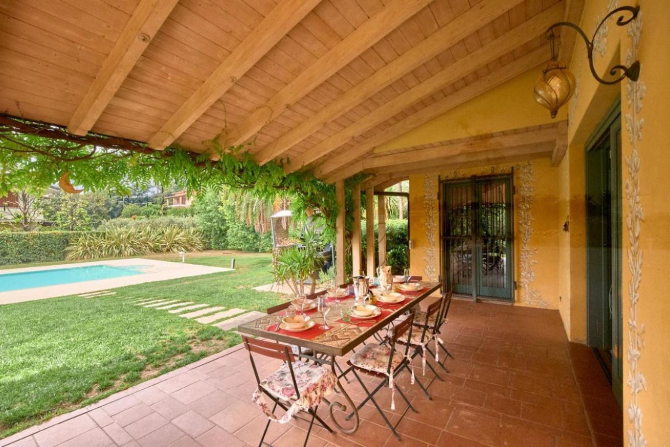 An outdoor dining area with a pool, covered patio, and lush greenery.