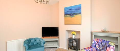A living room with a television, a blue armchair, a colorful armchair, a small table with a vase of flowers, and a painting on the wall.