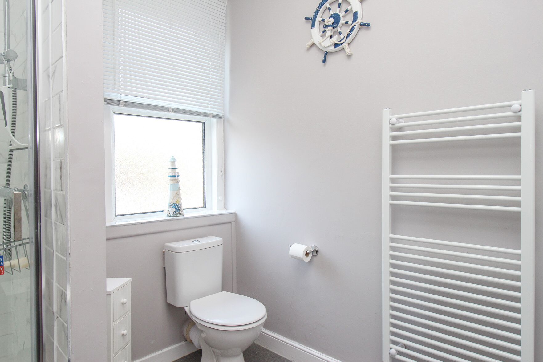A bathroom with a toilet, a window with a view, a radiator, and a ship wheel decoration.
