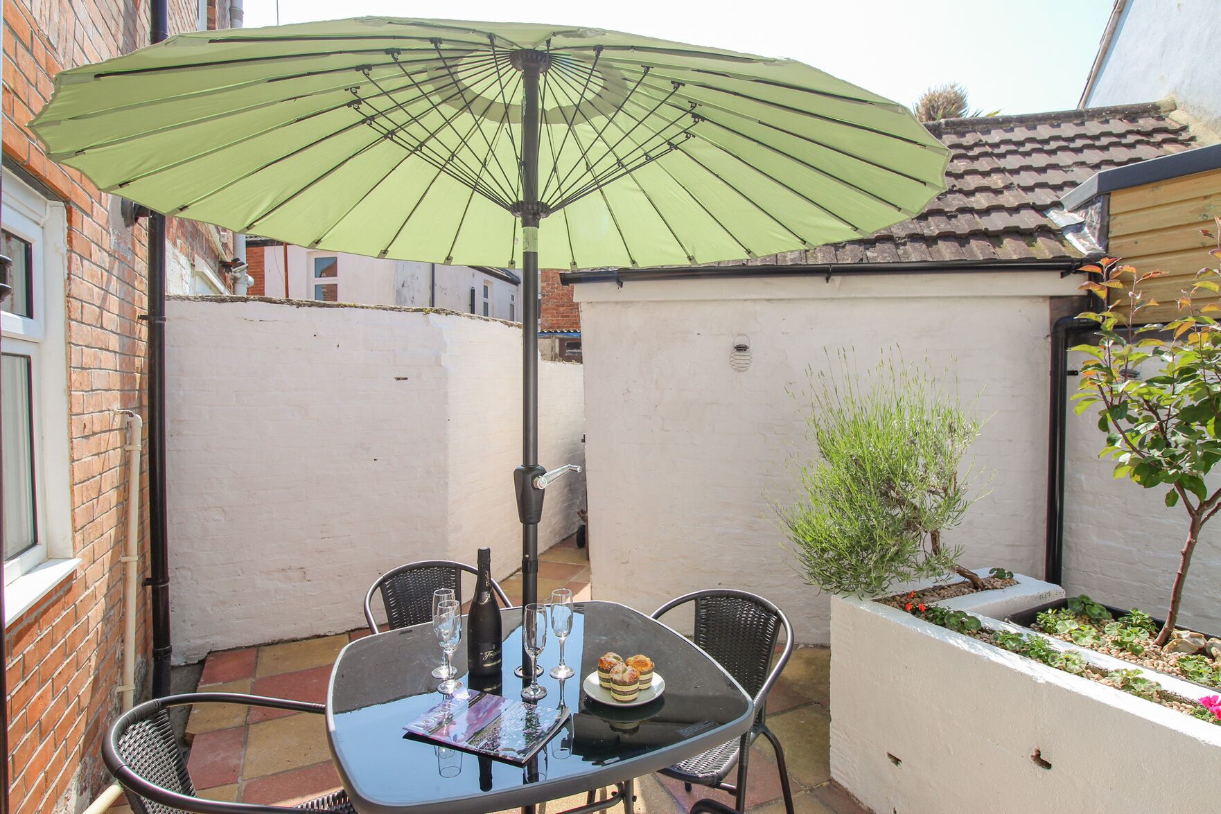 Eine Terrasse mit Tisch, Stühlen, einem grünen Sonnenschirm und einem kleinen Garten.