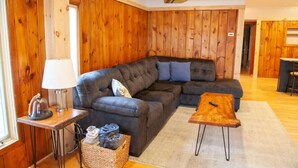 A cozy living room with a sectional sofa, a small wooden coffee table, a lamp, and a basket of items. - Lakefront Cottage: Dock & Fire Pit- Pet Friendly (Geneva)