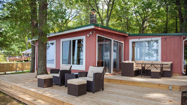 A covered deck with outdoor furniture, a sliding glass door, and a red building with a chimney. - Lakefront Cottage: Dock & Fire Pit- Pet Friendly (Geneva)