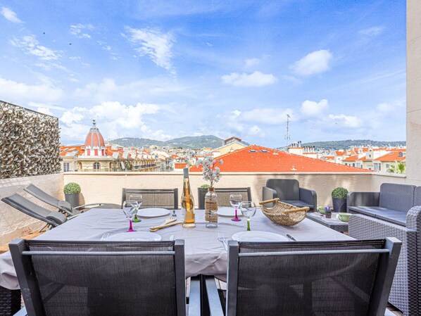 A rooftop dining area with a table set for two, overlooking a cityscape with red-roofed buildings and a dome structure.