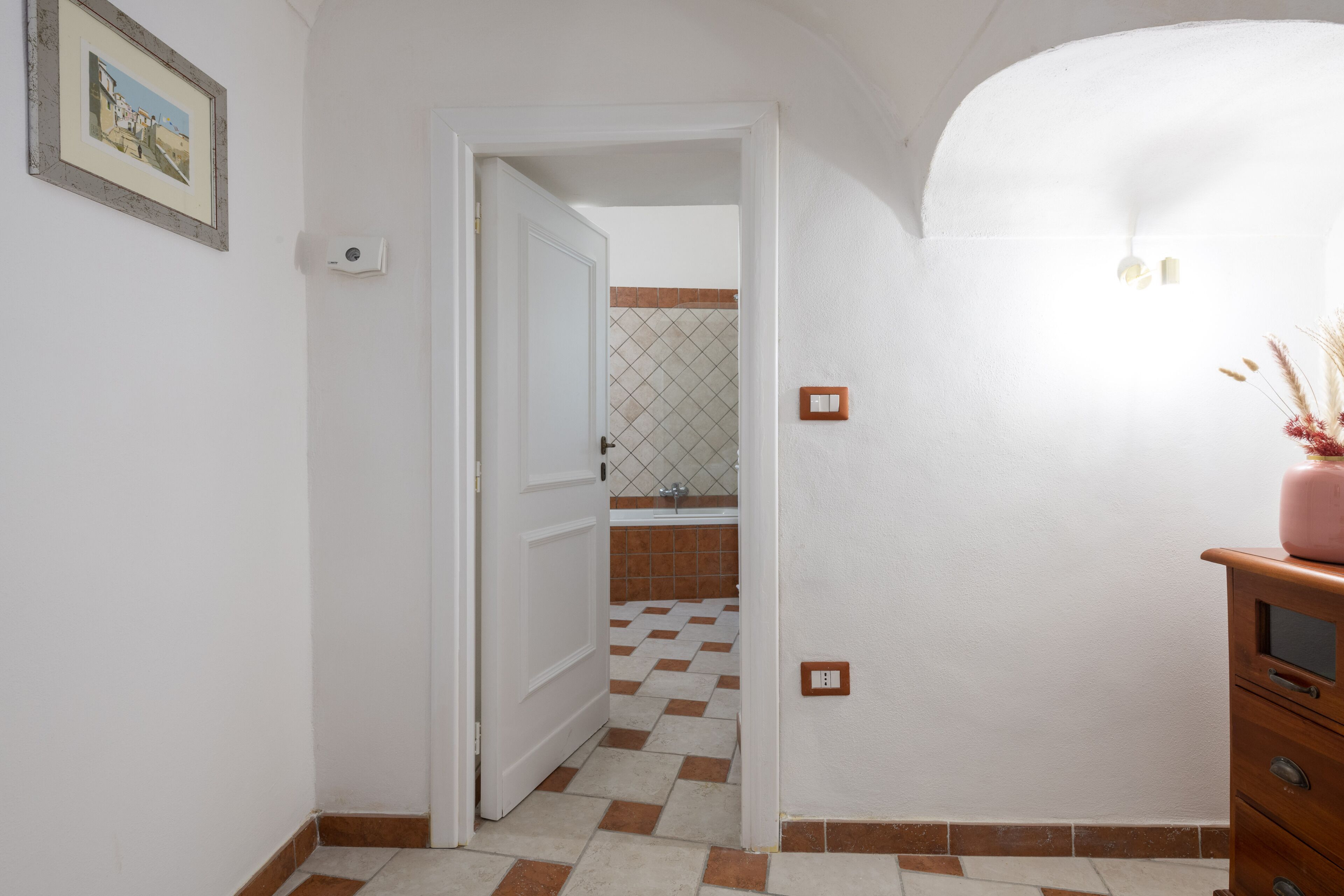 Un corridoio con pavimento a scacchi, una porta bianca che dà su un bagno e un settimino di legno con un vaso di fiori secchi.