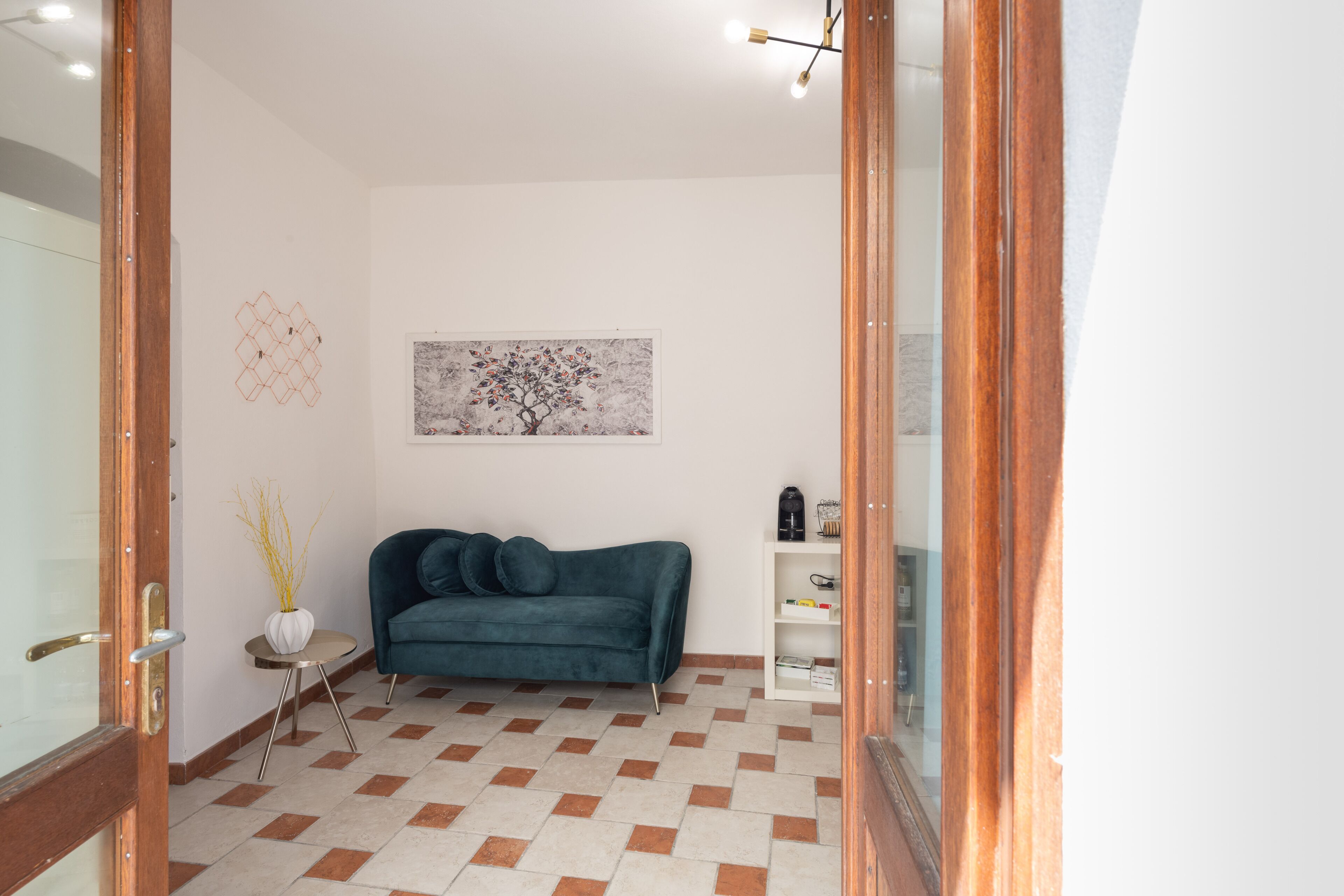A room with a teal sofa, a small side table, a vase with branches, a painting of a tree, and a white shelving unit.