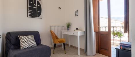 Una habitación con un sofá gris, un escritorio blanco, una silla y una puerta que da a un balcón.